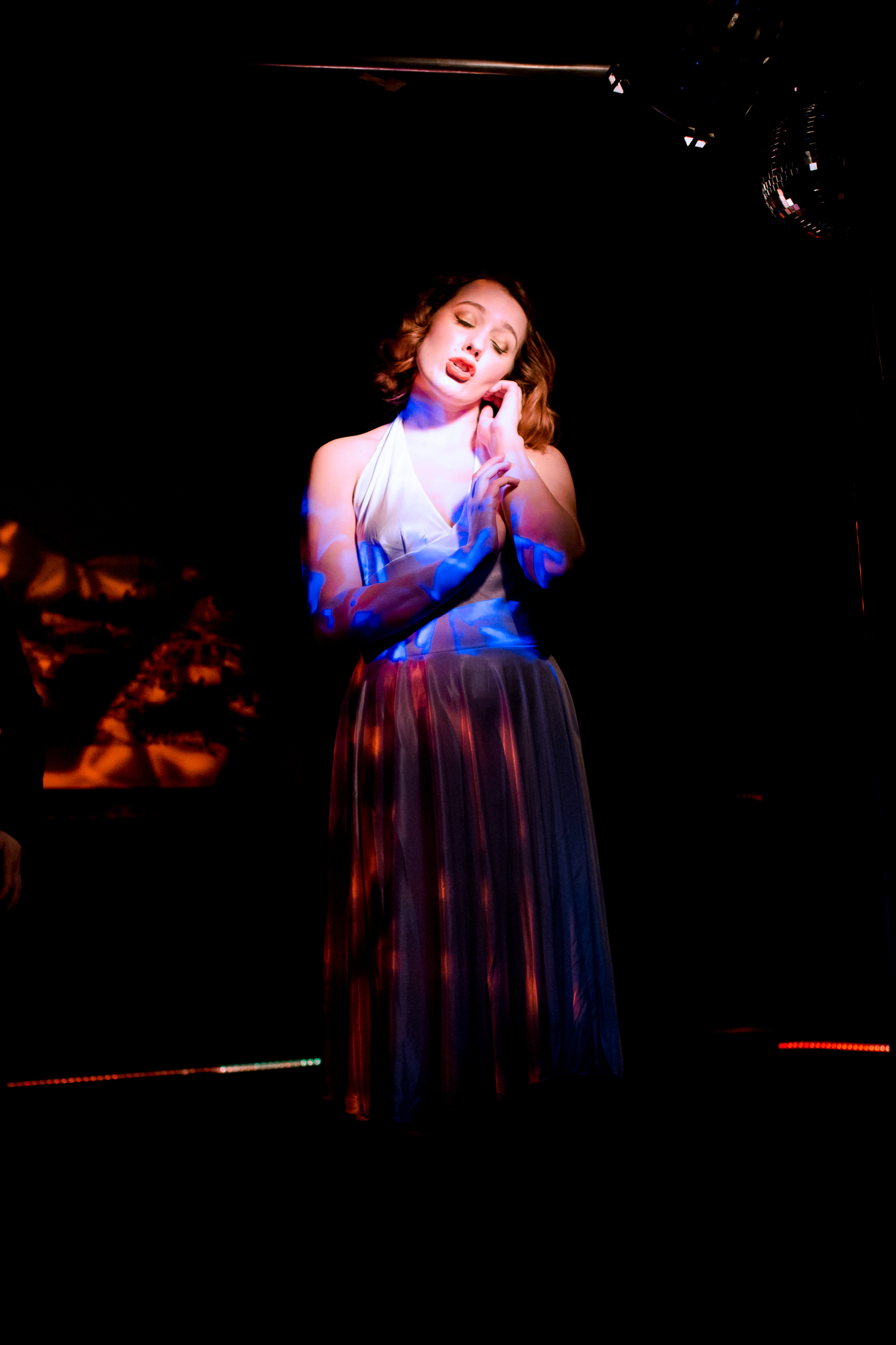 Blue Monday is a young white woman with shoulder-length brown hair. She is dressed in the classic Marilyn Monroe white halterneck dress. She ihas her eyes closed and is holding her arms close to her body, one hand caressing her cheek in a self-soothing manner. She is lit with red dappled light.