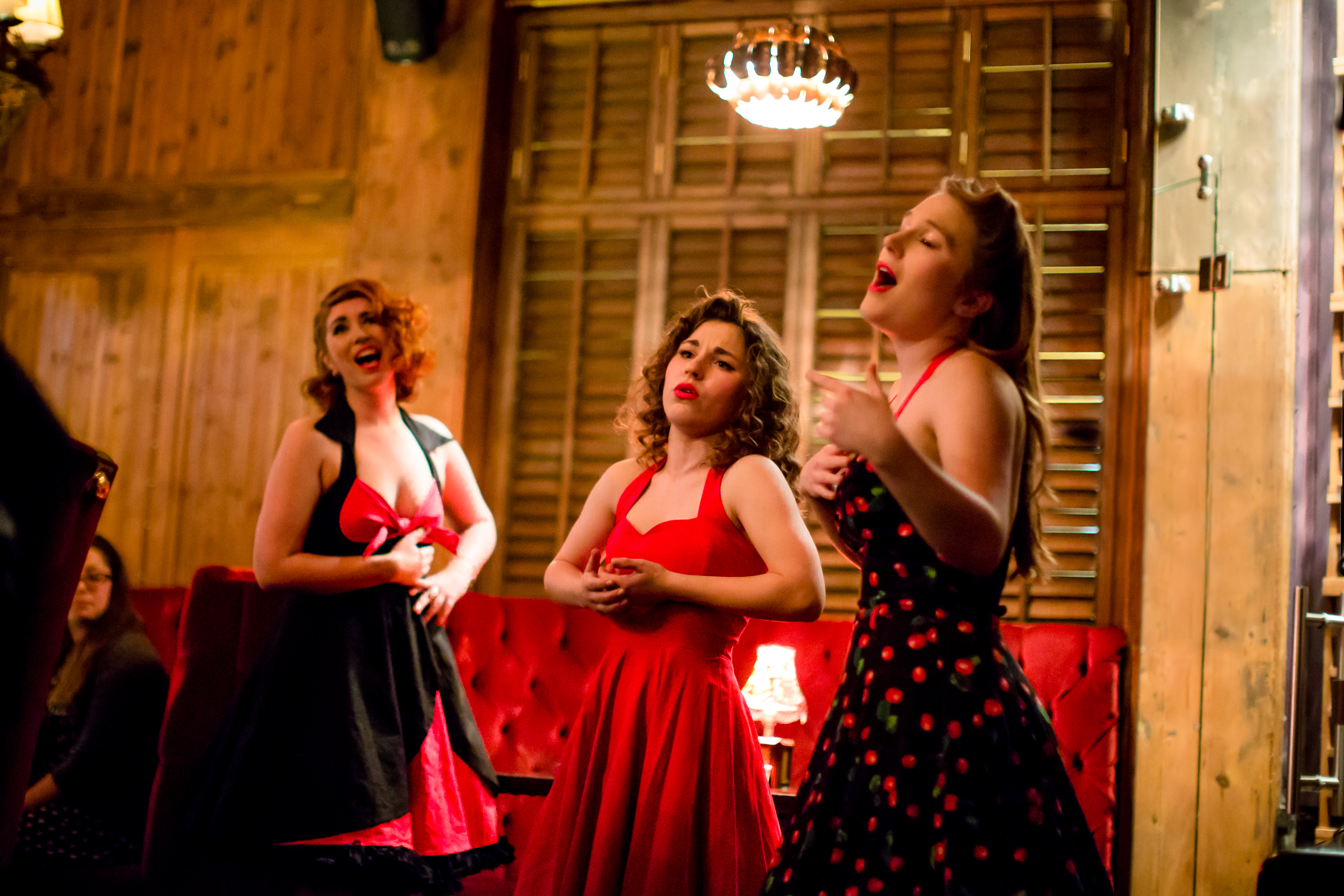 Three young white women, dressed in red and black 50's skater dresses of differing styles. They are all singing. Behind them are the wooden structures and plush interiors of Kings Head Private Members Club.