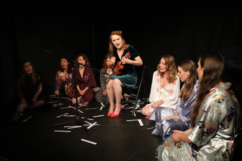 Ferrero Rochelle sits in the centre with a ukulele, she is singing. The troupe sit in a semi-circle at her feet, dressed in silk dressing gowns, also singing.