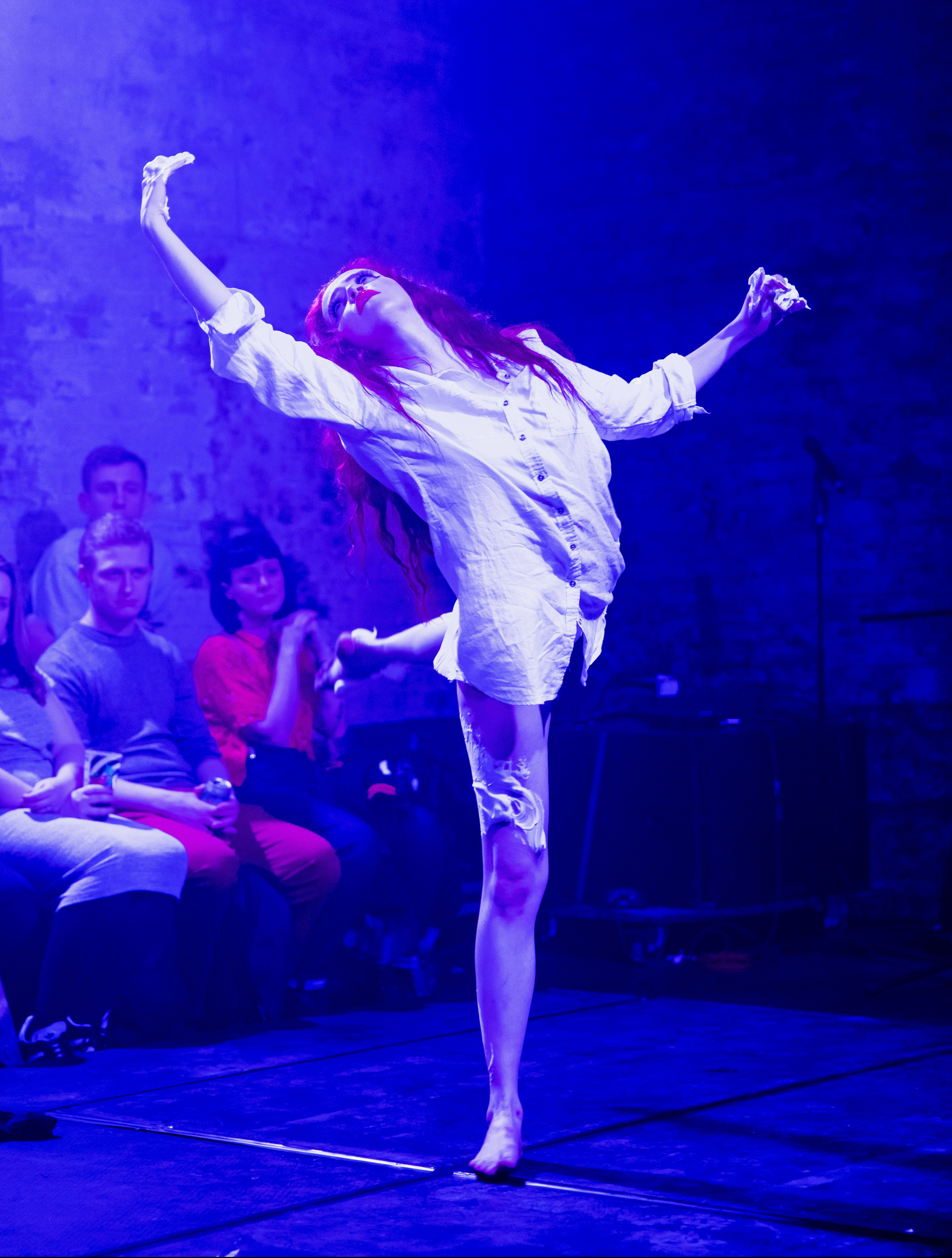 Ida Sanguine is a burlesque performer and classically trained dancer. She is mid-performance, in a glorious arabesque position. She is wearing a white evening-type shirt that covers her hips, and her legs are bare. Her long red hair falls down her back. Behind her we can see a few audience members, concentrating on her, entranced.