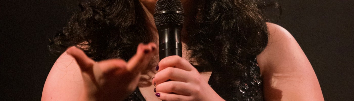 Rosie Verbose is a singer, comedian and MC. She is making a funny Grumpy Cat face, holding the microphone up with her left hand and stretching her other arm out. She is wearing a black, vintage-style, shoulder-length wig and a headdress made of four bright, colourful pom-poms. She also wears a black sequinned evening dress and a brightly coloured shawl, with stripes the same colours as the pom poms.