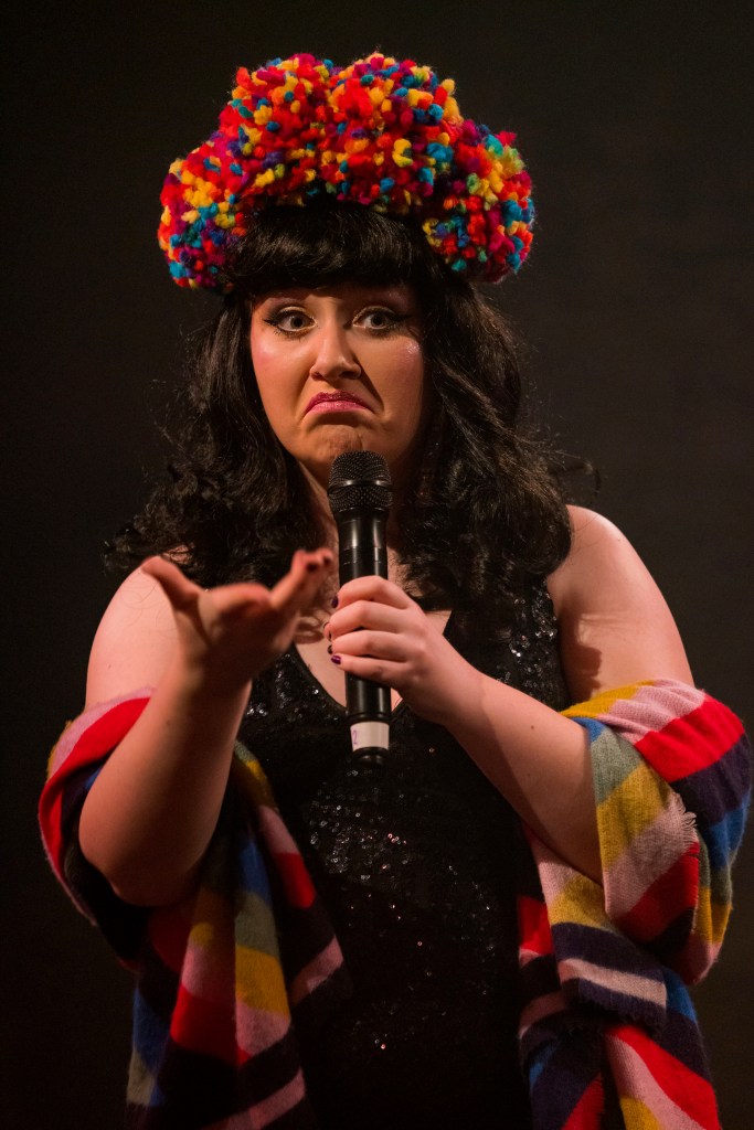 Rosie Verbose is a singer, comedian and MC. She is making a funny Grumpy Cat face, holding the microphone up with her left hand and stretching her other arm out. She is wearing a black, vintage-style, shoulder-length wig and a headdress made of four bright, colourful pom-poms. She also wears a black sequinned evening dress and a brightly coloured shawl, with stripes the same colours as the pom poms.
