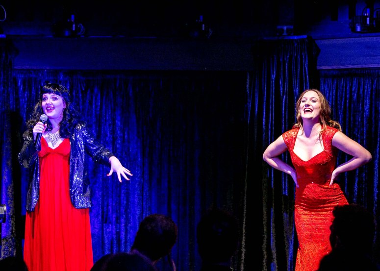 Performers Rosie Verbose and Ferrero Rochelle, hosts of this mental health podcast, stand on opposite ends of the stage, both dressed in scarlet. Rosie is also wearing a black sequin blazer and a 50s-style black wig. She is holding a microphone and is saying something animatedly. Rochelle is laughing. Her auburn hair is down and her hands are on her hips.