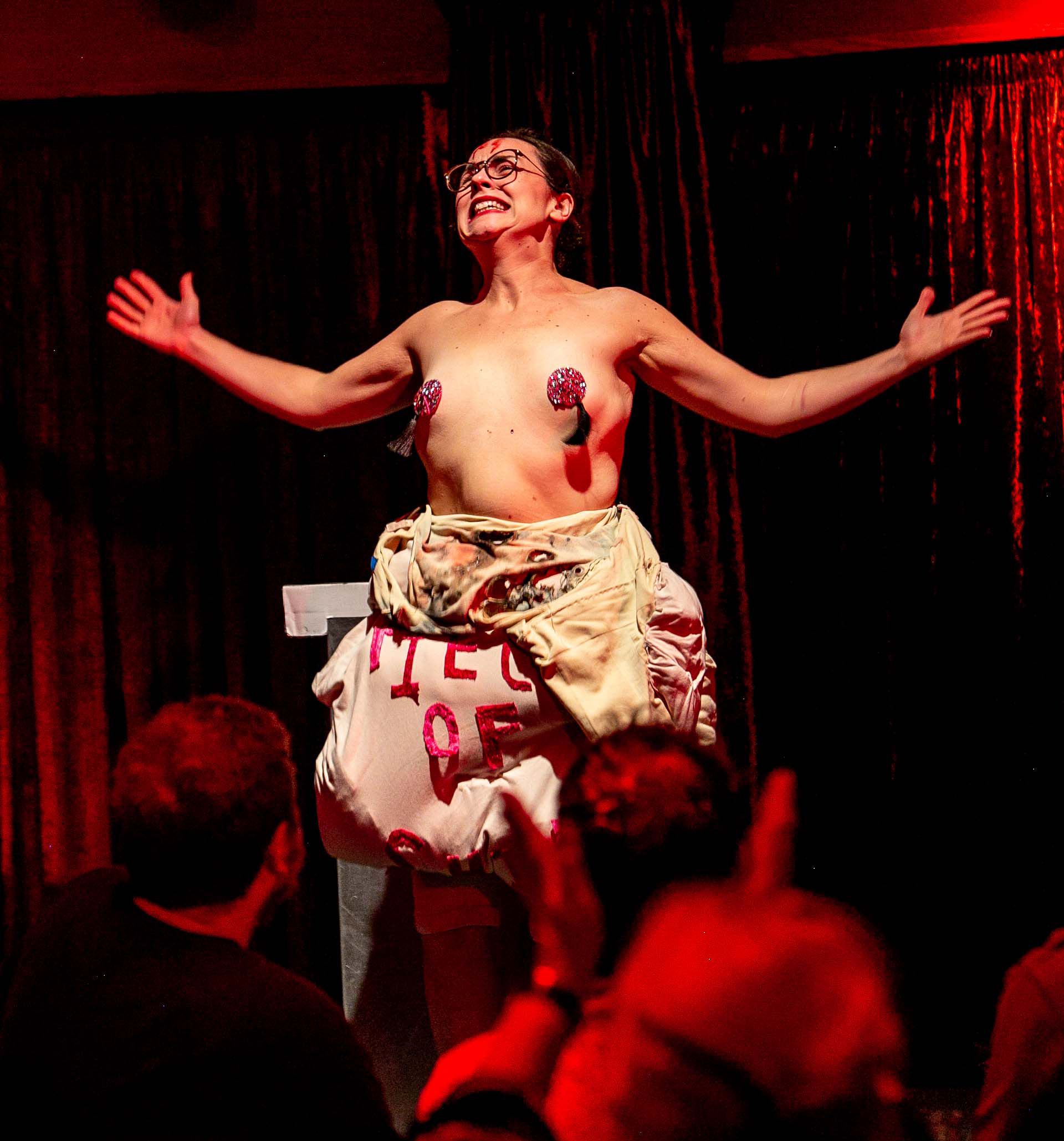 Mouncealot performing her act about C-PTSD at an Invisible Cabaret show. She is stood on stage with her arms flung wide, wearing a pair of red pasties and is halfway through removing a cumbersome bodysuit.