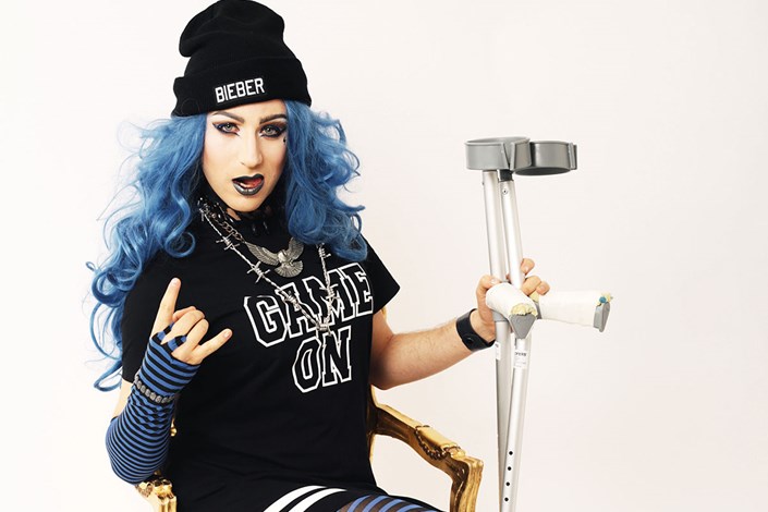 Guy dressed as his alter ego, Electra, wearing a blue wig, holding a crutch in one hand and making the 'rock' sign with the other