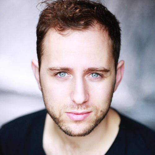 Professional headshot of Guy Woolf, who has blue eyes, wavy brown hair and stubble. He is wearing a plain black T-shirt and looking enigmatically into the camera.