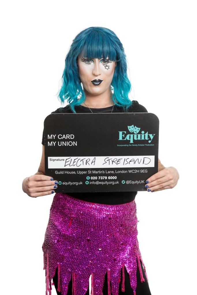 Guy is dressed as his drag queen alter ego, Electra. He is wearing a bright blue bobbed wig, elaborate black eyeliner with a Star of David on his right cheek, black lipstick and dark blue nail polish. He is wearing a black T-shirt and bright pink sequin wrap skirt over black leggings. She is holding an oversized Equity card in front of her. The card says 'Equity: My Card, My Union' and is signed 'Electra Streisand.' 