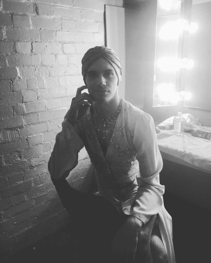 A black and white photo of Mateo Oxley as Prior Walter in drag. He has his back to the dressing room mirror, which is lit with large bulbs in that classic ‘dressing room mirror’ way. He is wearing a long-sleeved vintage-style dress with a 1930s-style turban and a bejewelled necklace. He is resting one hand lightly on his cheek, leaning on the back of his chair.