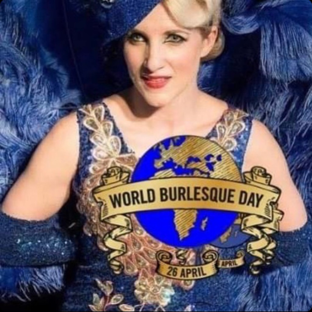 Sapphira is a blonde showgirl dressed in royal blue. She is holding giant blue ostrich feather fans behind her. On top of this image is the World Burlesque Day logo, a globe with a banner across and underneath it.