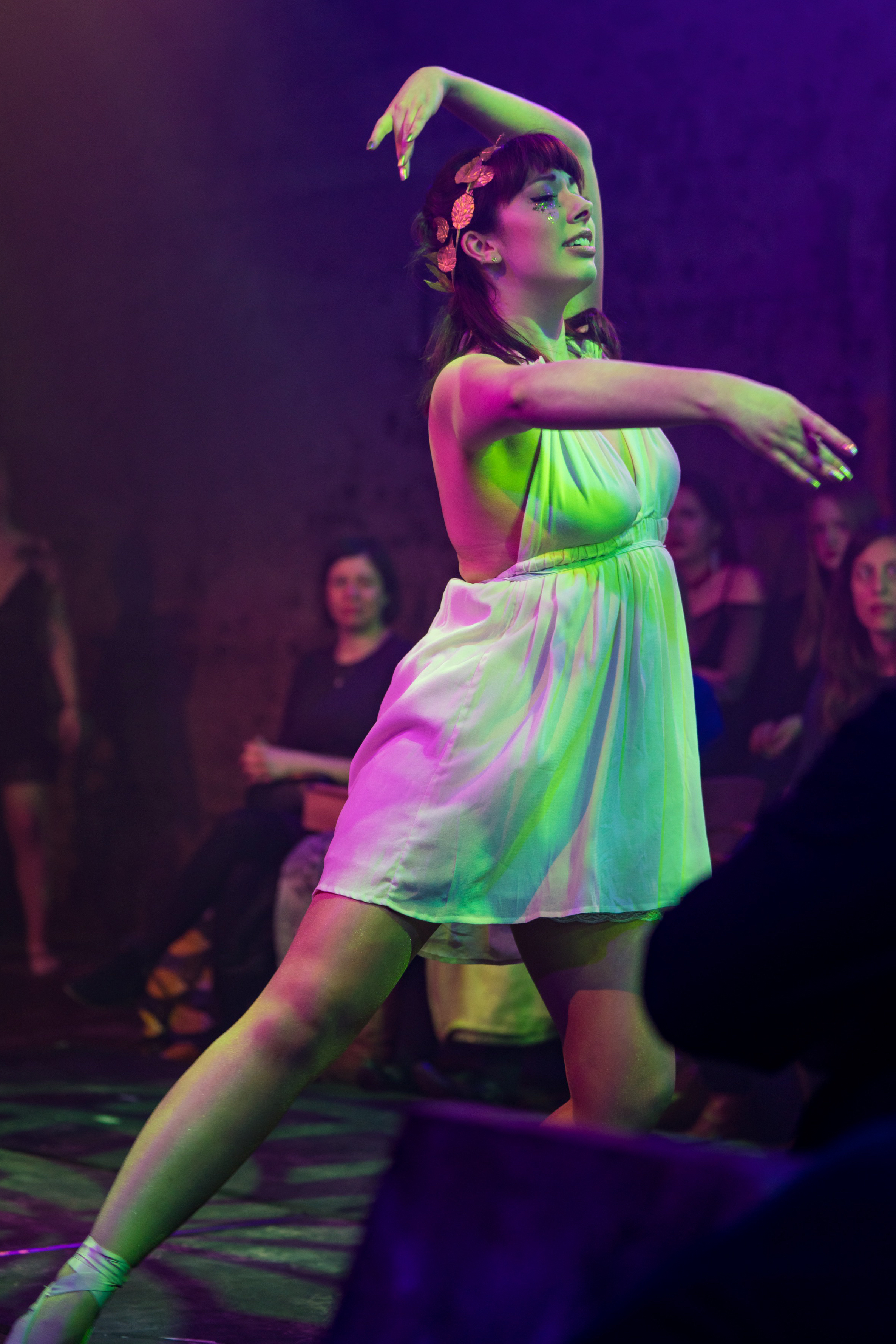 Belle Bluestocking is a dancer and cabaret performer. She is mid-dance here with her arms in fourth position, and one leg extended to the side. She is wearing a white, flowing, Grecian-looking mini-dress, and a gold leaf crown. She has collar-length brown hair. Her eyes are closed and she looks focused and serene. Behind her are several audience members watching on.