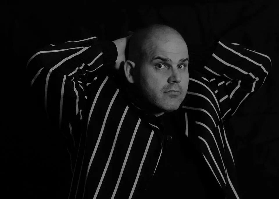 A black and white photo of Richard 'The Burly Photographer', a bald man dressed in a striped shirt, who is passionate about men's mental health. He is posed with his hands behind his head, looking interrogatively at the camera.