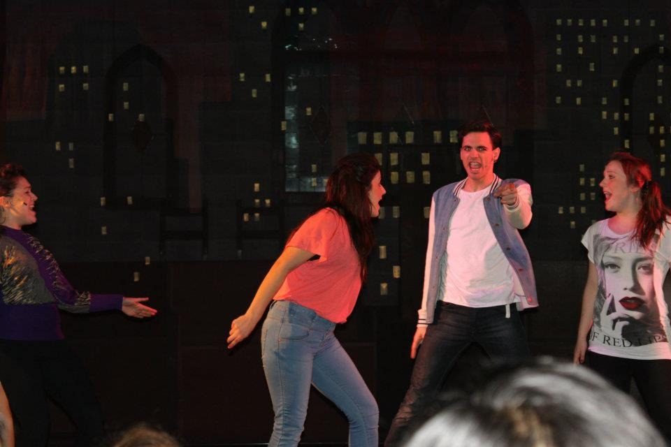 Four young people onstage in a musical theatre dance number. Mateo is wearing a grey jacket with white sleeves and jeans. He is pointing into the audience and singing. To his left on the righthand side of the frame is Rochelle, wearing her auburn hair in a ponytail and a band T-shirt. She is looking at Mateo and also singing.