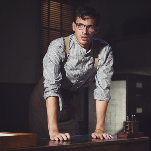 Mateo Oxley onstage, in character, dressed in a striped blue shirt with the sleeves rolled up, brown trousers, braces and glasses. He is leaning on a desk and looking concerned.