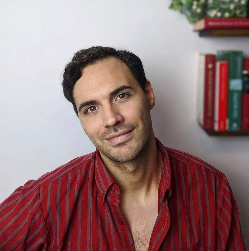 Mateo Oxley, a young man with brown eyes, dark brown hair and a hint of stubble, poses for a relaxed headshot. He is wearing a striped red shirt with the top buttons undone. He is leaning on a white wall, on which we see the hint of a bookshelf and houseplant.